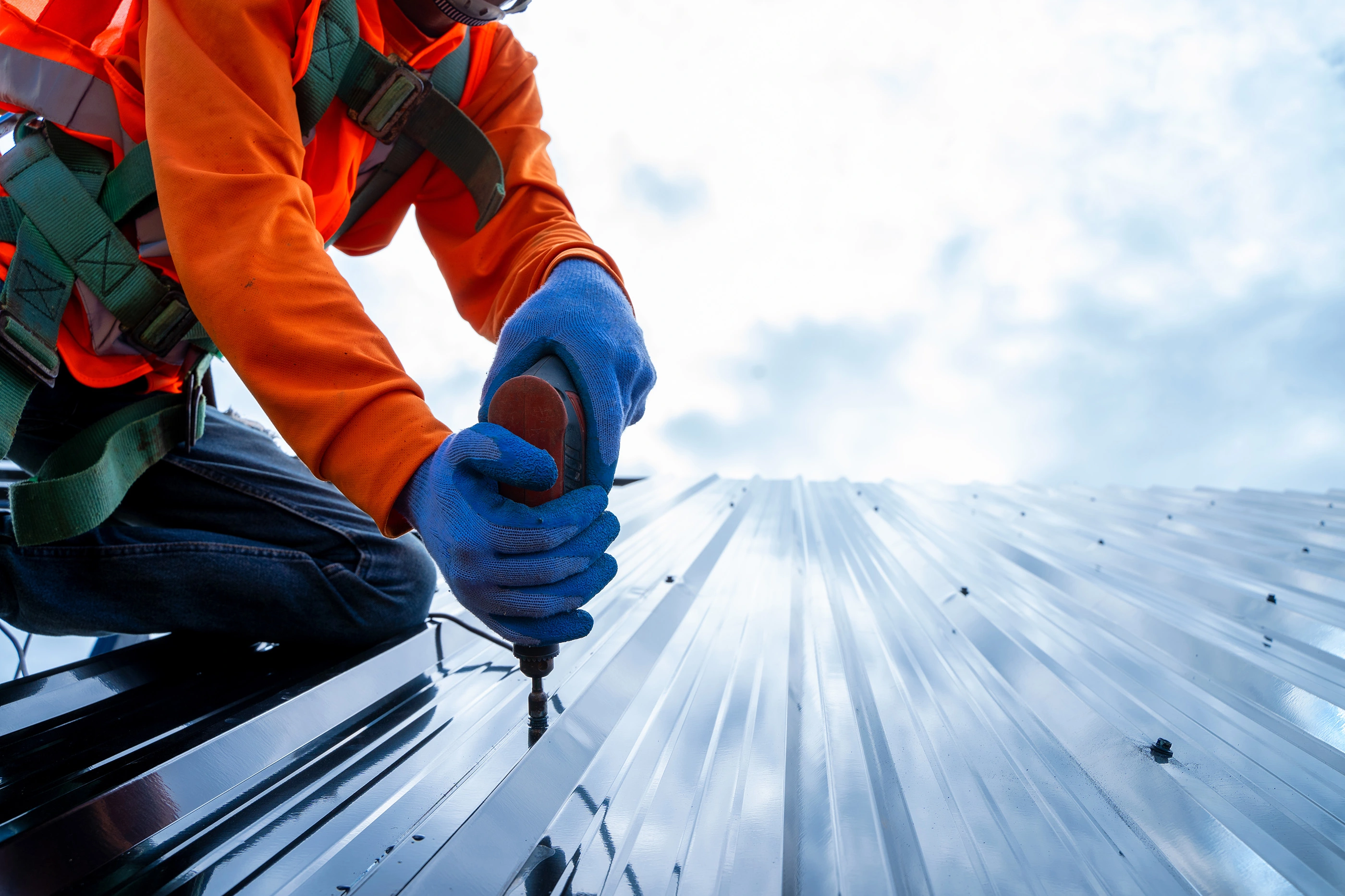 Closeup of person in orange hi-vis jacket wearing harness and blue gloves using a drill on top of a roof