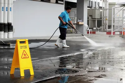 Photo taken of a man using a high pressure water jet, wearing white boots next to a caution sign.